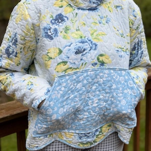 May include: A quilted hoodie in a floral pattern featuring blue, yellow, and white flowers on a light blue background. The hoodie has a kangaroo pocket and a hood. The garment is worn over checkered pants.