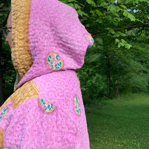 Puede incluir: Chaqueta acolchada rosa con capucha. La chaqueta tiene un forro y ribete con estampado amarillo y dorado. La tela rosa est&aacute; decorada con dise&ntilde;os florales bordados. La chaqueta se usa al aire libre con un fondo verde.