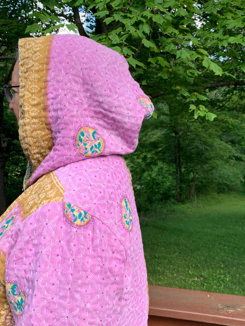 Puede incluir: Chaqueta acolchada rosa con capucha. La chaqueta tiene un forro y ribete con estampado amarillo y dorado. La tela rosa est&aacute; decorada con dise&ntilde;os florales bordados. La chaqueta se usa al aire libre con un fondo verde.