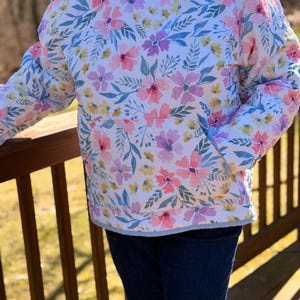 May include: A quilted hoodie with a floral pattern in pink, purple, and yellow on a white background. The hoodie has a kangaroo pocket and a light blue trim. The sleeves are long and the hood is lined with a striped fabric.