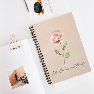 Puede incluir: Un cuaderno de espiral con una cubierta beige que presenta una ilustración de acuarela de una rosa rosa con hojas verdes. El texto "Truth. Goodness. and Beauty." está escrito en cursiva debajo de la rosa.