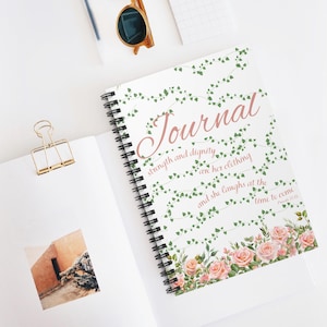 Puede incluir: Un diario blanco con un diseño floral y el texto "Journal" en rosa. La cita "Fuerza y dignidad son su vestimenta, y ella se ríe en el momento de venir" está impresa en rosa con la referencia "Proverbios 31:25".