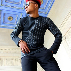 May include: A man wearing a black long-sleeved shirt with a quilted design on the left side. He is wearing black pants and sunglasses. The background is a white wall with a blue and white tiled ceiling.