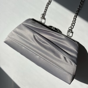 May include: A silver-gray clutch with a chain strap. The rectangular bag has a pleated design on the front and a silver clasp. The chain strap is also silver. The bag is made of a shiny, satin-like material.