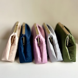 May include: Five rectangular clutch bags with gold-toned metal kiss-lock frames, arranged in a row. The bags feature a soft, satin-like fabric in shades of pale pink, navy blue, light purple, silver-grey, and olive green with gathered side details.