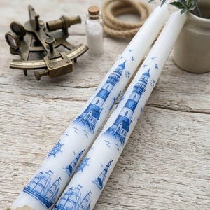 May include: Two white taper candles decorated with blue lighthouses and sailboats. Nautical elements include a sextant, rope, and a small jar of salt. The candles have the text "Hand-Made Coastal Tapers" on a tag.