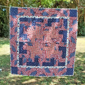 May include: A square quilt featuring a patriotic design with a central block of red and white stripes surrounded by a border of blue fabric with white stars. The pattern repeats the American flag motif throughout the entire textile surface.