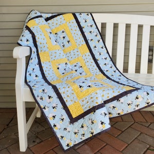 May include: A blue and yellow quilt with a black border and a pattern of sheep and bees. The quilt is folded and draped over a white bench.