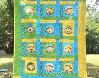 May include: A patchwork quilt featuring twelve circular panels, each displaying a cartoon monkey with a unique facial expression. The quilt is constructed with alternating blue, green, and yellow fabric squares, finished with a yellow border.