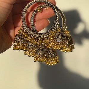 May include: Gold-coloured hoop earrings with bell-shaped charms. The earrings have detailed filigree patterns and small, dangling beads. The design is similar to traditional Indian jewellery, with a vintage appearance.