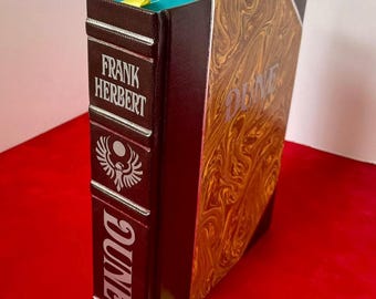 May include: A brown leather-bound book with a gold-colored title, "Dune", and the author's name, "Frank Herbert", on the spine. The book has a yellow bookmark and a teal-colored top edge.