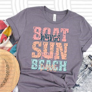 May include: A gray t-shirt with the words "Boat Waves Sun Beach" in a distressed, colorful font. The shirt is paired with denim shorts, sunglasses, a straw hat, and a colorful scarf, suggesting a beach theme.