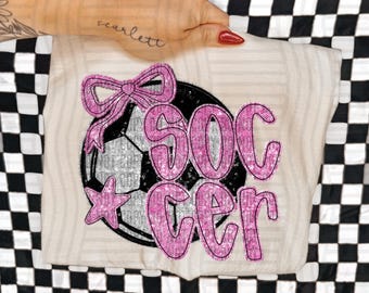 Könnte beinhalten: Beiges T-Shirt mit einem Fußball-Design. Der Fußball ist schwarz und silber, mit einer rosa Schleife und einem Stern. Das Wort "SOCCER" ist in rosa Glitzerbuchstaben geschrieben.