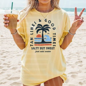 May include: A light yellow t-shirt with the text "TAN LINES & GOOD TIMES" and "SALTY BUT SWEET just add water" in black and blue. The graphic includes a palm tree, sun, and ocean scene. The shirt is worn on a beach.