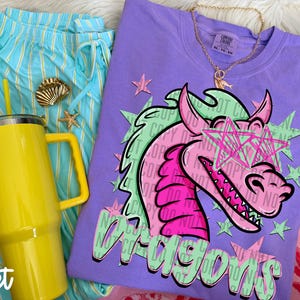May include: A violet t-shirt featuring a graphic of a pink dragon wearing star-shaped glasses, surrounded by light green stars and the word Dragons in light green text. A yellow tumbler with a handle and a gold chain necklace are also visible.