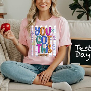 May include: A light pink t-shirt with the words "YOU GOT THIS" in colourful, playful lettering. The shirt has a grid and star design. A small chalkboard with the words "Test Day" is visible.