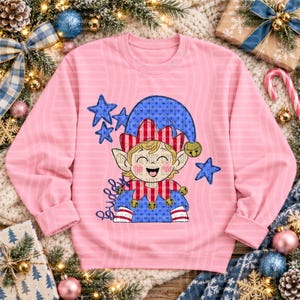 Op de afbeelding: Een lichtroze sweatshirt met lange mouwen en een afbeelding van een elf met een blauw-rood gestreepte muts en kraag. De elf heeft puntige oren, een lachend gezicht en kleine belletjes. Blauwe sterren omringen het figuur en het woord Buddy staat in blauwe schrijfletters aan de linkerkant.