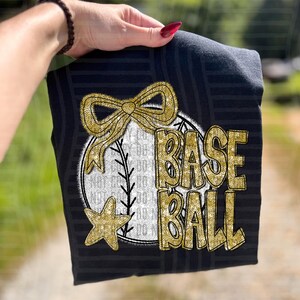 May include: Black t-shirt with a gold glitter baseball design. The design includes a baseball, bow, and star, with the word "BASEBALL" in gold glitter lettering. The shirt has a striped pattern.