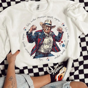 May include: White sweatshirt featuring a graphic of a man in a cowboy hat and a jacket with stars and stripes. The text "Let's make America great again" is written around the image. The person is wearing blue jeans and black shoes.