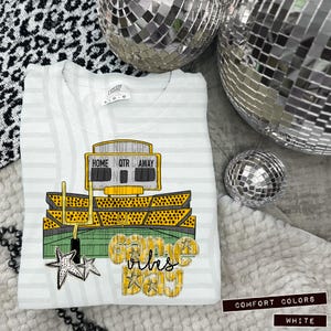 Puede incluir: Sudadera blanca Comfort Colors con un gráfico de estadio de fútbol. El diseño incluye un marcador, postes de portería y las palabras "Home Qtr Away" y "Game Day Vibes". La sudadera tiene un patrón de rayas.