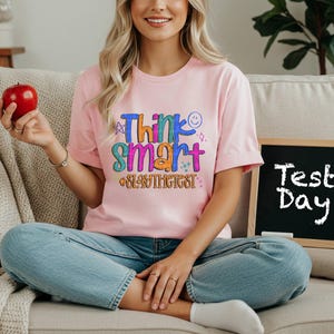 May include: A light pink t-shirt with the words "Think Smart #SlayTheTest" in colorful text. A small chalkboard with the words "Test Day" is visible. The person is holding a red apple.