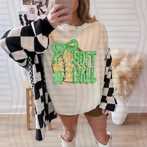 May include: Cream-colored sweatshirt with a softball graphic. The graphic features a gold softball with a green bow and the words "SOFT BALL" in green. The person is wearing a black and white checkered cardigan and a baseball cap.