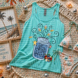 Op de afbeelding: Een turquoise tanktop met een afbeelding van een weckpot met de tekst "Let Your Light Shine" en vuurvliegjes. De afbeelding bevat ook een bikini set met bloemenprint en ingelijste strandkunst.