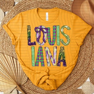 May include: Mustard yellow t-shirt with the word "LOUISIANA" in a colourful, patterned design. The letters are decorated with purple, green, and gold patterns, including leopard print and plaid. The shirt is knotted at the bottom.