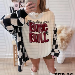 May include: Cream-colored sweatshirt with the text "let's play some BASE BALL" in red and black. The sleeves are black and white checkered. A baseball cap and white boots complete the look.