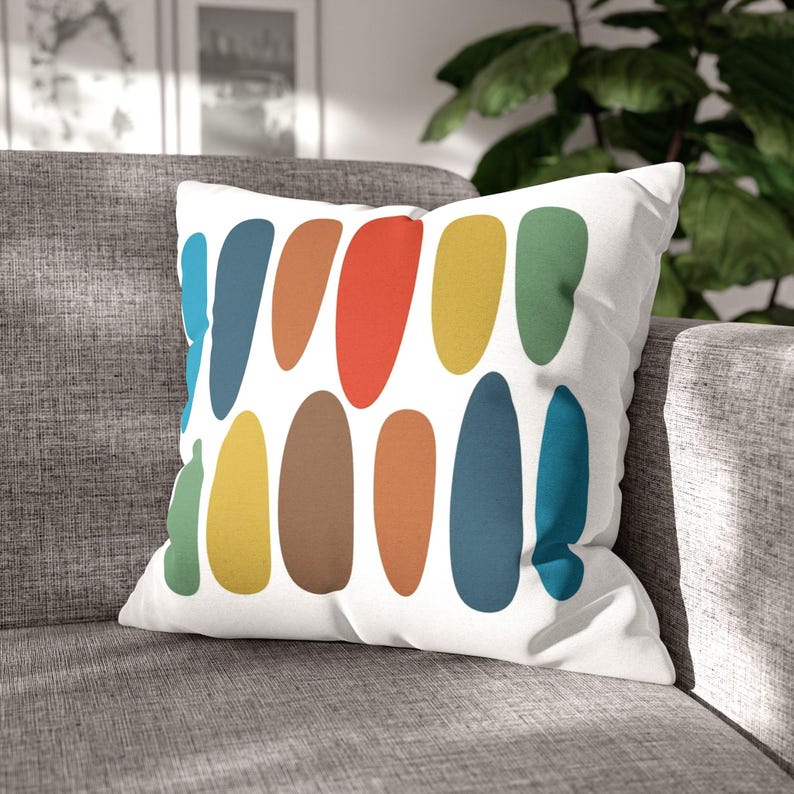 Mid-century modern pillow with colorful oval pattern resting on a gray sofa, with sunlight & a leafy plant in the background.