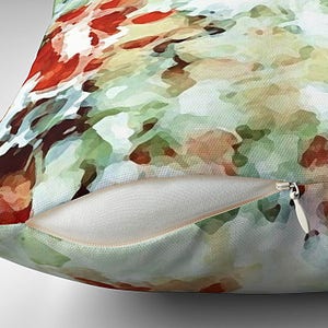The pillow in the image is a square throw pillow with an abstract floral design that features washes of green, red, & brown watercolor paint. The pillow cover is partially unzipped to show the hidden zipper and insert.
