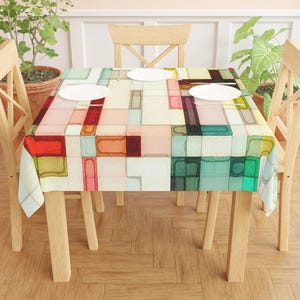 May include: A rectangular tablecloth with an abstract geometric pattern in a variety of colors, including red, green, pink, and yellow. The design features a grid of rectangular shapes. The tablecloth is on a wooden table with four chairs and white plates.