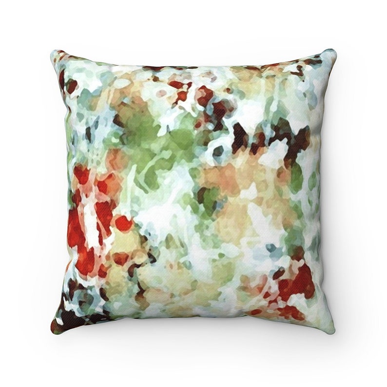 A plump throw pillow with heavily abstracted, softly focused flowers and leaves appear to be growing in a lush, untamed garden. The watercolor design is in shades of sage green and reddish light brown with spots of vivid red and white.