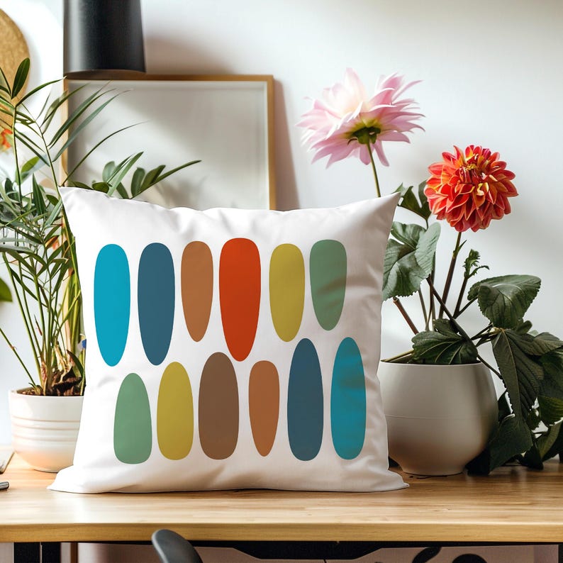 White pillow with colorful mid-century oval pattern on a wood desk, surrounded by potted plants & bright flowers.
