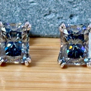 May include: A pair of square-cut, gray gemstone stud earrings. Each earring features a large, faceted stone set in a silver-colored metal with decorative prongs. The earrings are displayed on a wooden surface.