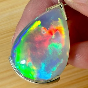 May include: A pear-shaped opal pendant with a silver setting. The opal displays a vibrant play of color, including green, yellow, red, and blue hues. The iridescent gemstone is suspended from a silver bail, creating a striking piece of jewelry.