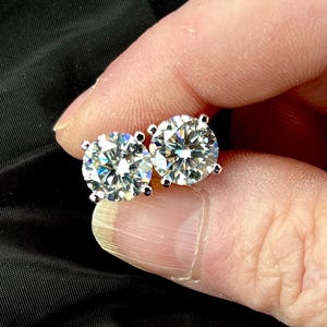 May include: A pair of sparkling, round-cut diamond stud earrings. The diamonds are set in a silver or platinum setting, and they are held between fingers against a dark background. The earrings are a classic jewelry item.