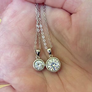 May include: Two silver necklaces with round, clear gemstone pendants. The pendants are set in a bezel setting and hang from delicate silver chains. The necklaces are displayed against a light background.