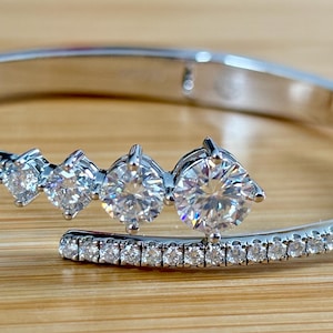 May include: A silver-toned cuff bracelet with a row of small, clear stones and a series of larger, clear, round-cut stones. The bracelet is set against a light brown wooden surface.