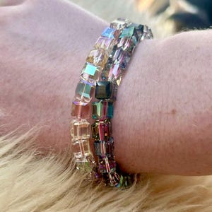 May include: Two bracelets made of iridescent square beads. The top bracelet is made of clear beads with a rainbow sheen. The bottom bracelet is made of dark purple, blue, and green beads with a rainbow sheen.