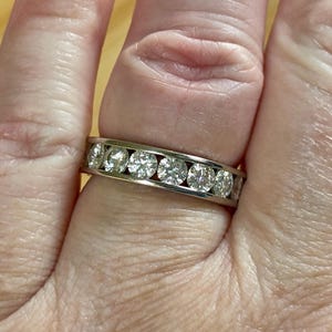 May include: A silver band ring with seven round, clear gemstones set in a channel. The ring is shown on a finger, with the gemstones reflecting light.