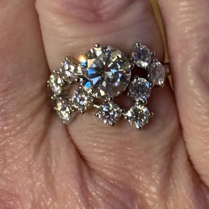 May include: A silver ring with a large round center stone and smaller round stones surrounding it. The ring is set on a finger.