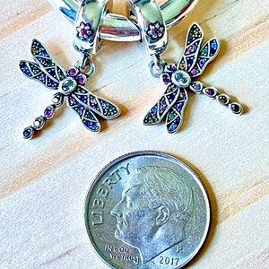 May include: Two silver dragonfly charms with colorful crystal accents. The charms are hanging from a silver hoop. A US dime is included for size reference.