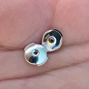 May include: Two silver-colored earring backs. Each back has a circular design with a central hole and a curved, notched edge. The text "S925" is visible on one of the backs. The backs are shown against a skin-toned background.