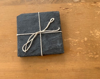 May include: A set of four black slate coasters tied together with twine. The coasters are square and have a rustic, natural look.