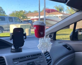 May include: A red and white crocheted coffee cup car air freshener hanging from the rearview mirror of a car. The air freshener has the text "You look good" on it.