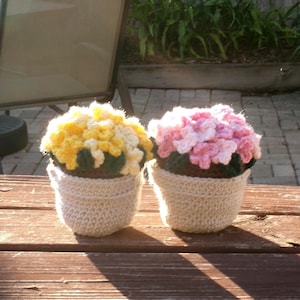Puede incluir: Dos macetas de flores de ganchillo, una con flores amarillas y blancas, la otra con flores rosas. Las macetas son de color crema y están sobre una superficie de madera. Las flores tienen hojas verdes.