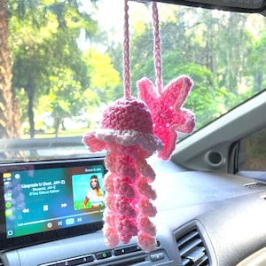 May include: A handmade, pink crochet jellyfish car charm with white accents and dangling tentacles. A matching pink starfish charm hangs nearby. The charms are suspended from a car's rearview mirror. The car's dashboard and a screen displaying music are visible.