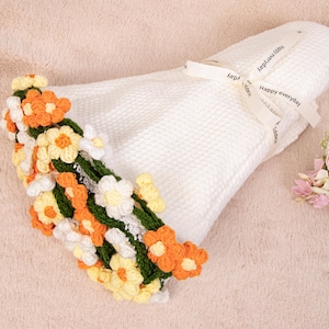 May include: A white waffle-textured fabric folded and tied with a cream ribbon printed with the text Happy everyday. One end is decorated with a cluster of crocheted flowers in shades of orange, yellow, and white with green stems and leaves.