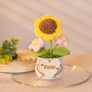 May include: A crocheted sunflower with pink flowers in a white heart-shaped pot with the word "Relax" on a wooden tag.